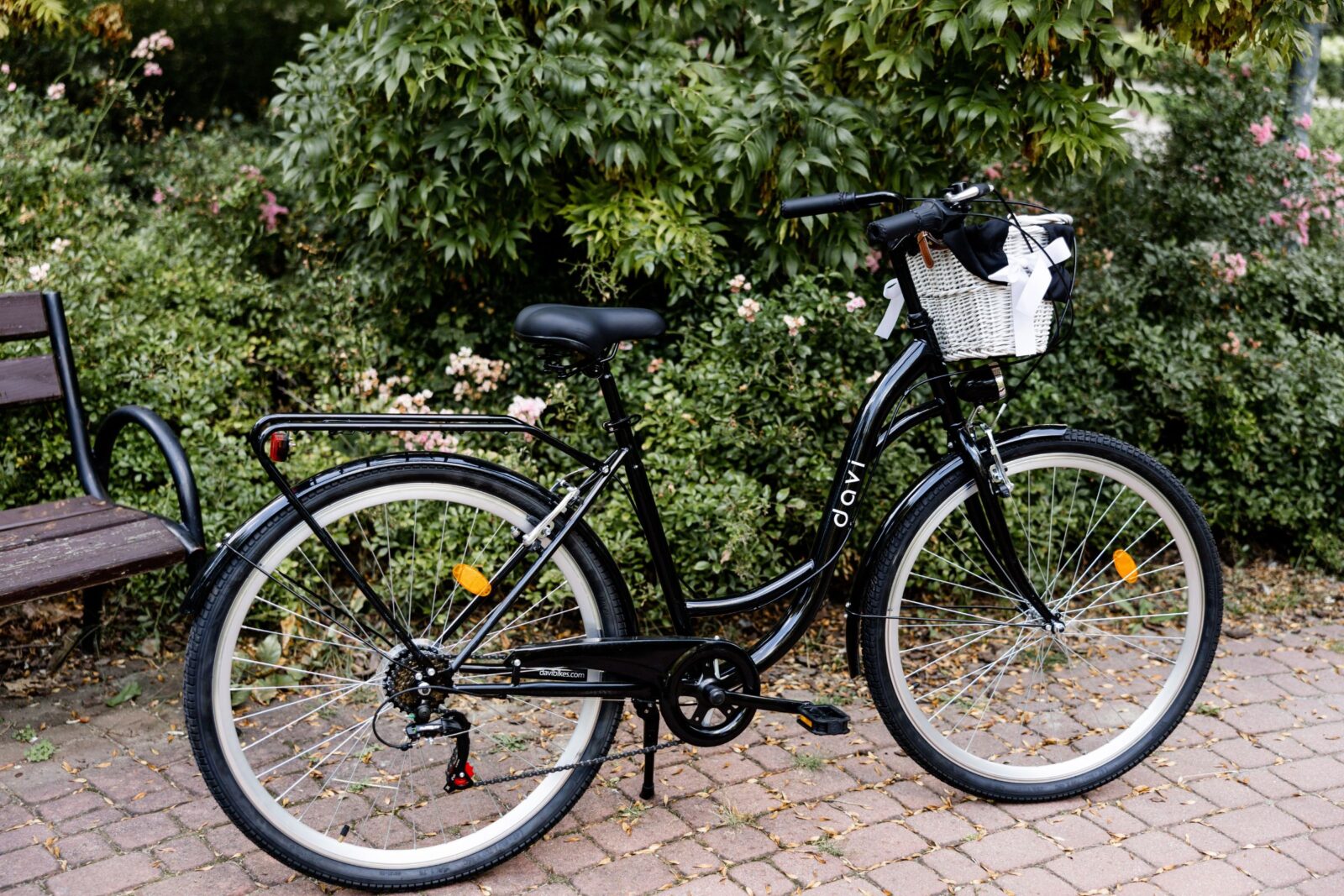 Svart cykel med korg vid parkbänk och buskar.